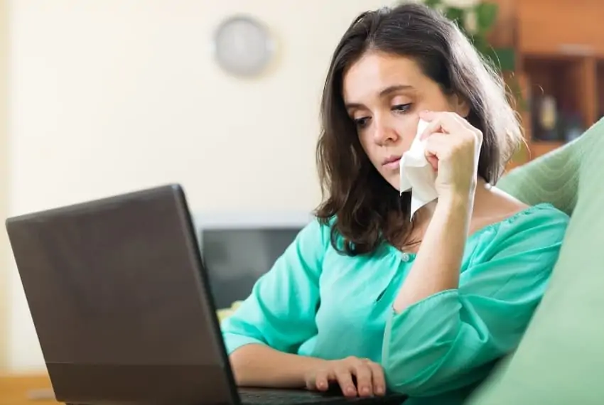 Woman looking at a laptop sadly while wiping her eye with a tissue.