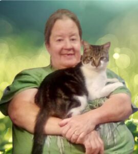 Person smiling while holding a tabby cat, with a serene outdoor backdrop blurred in the background.