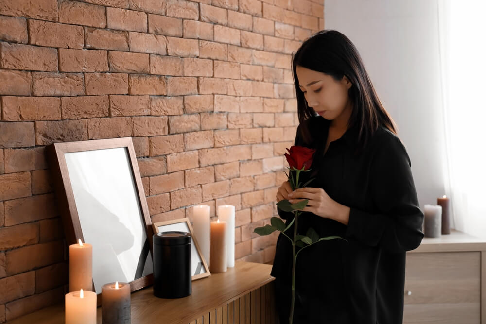 Woman Holding Red Rose by Candle-Adorned Table, Symbolizing Reflection on Life and Cremation, Brick Wall Background.