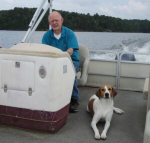 A man steering a boat on a serene lake, accompanied by a brown and white dog lounging on the deck; tranquil shoreline with lush trees under an overcast sky in Alabama - ideal for peaceful reflections on life, related to cremations, funerals, or pre-planning services.