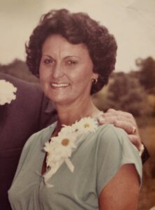 A woman in a light-colored dress with a floral corsage smiles outdoors; partial view of another person's hand on her shoulder. Ideal for depicting warmth and connection at Alabama Funeral Homes' cremations, funerals, and pre-planning services.