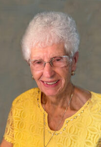 Elderly woman with white hair and glasses, wearing a yellow lace top, smiling gently against a neutral backdrop; highlighting warmth and tranquility in funeral and cremation services in Alabama.