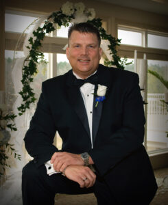 A man in a tuxedo at an Alabama funeral home stands before a floral arch, symbolizing funerals and cremation services.