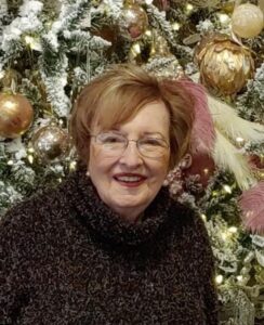 An older woman with glasses smiles by a Christmas tree, featured in our Alabama funeral home cremation and burial services.