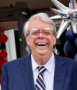 An older man in a blue suit smiles at a funeral home event in Alabama, with balloons and cremation services details.