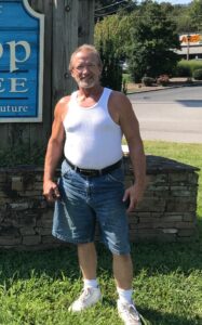 An older man stands on grass near a funeral home in Alabama, with a blue sign for cremation services and burial options.